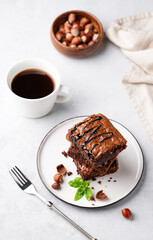 A stack of chocolate brownies with mint, hazelnuts and salted caramel on a white plate on a light background with cup of coffee and bowl with nuts.