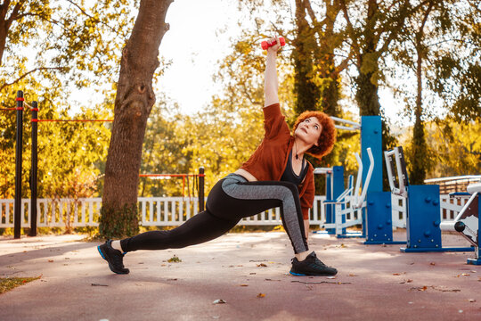 Caucasian pretty mid adult woman with ginger hair stretching using dumbbells. Low angle view. Fitness in city park