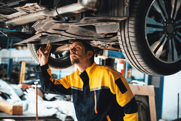 Vehicle mechanic conduct car inspection from beneath lifted vehicle. Automotive service technician in uniform carefully diagnosing and checking car's axles and undercarriage components. Oxus - obrazy, fototapety, plakaty