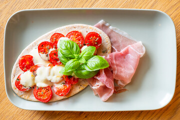 Tomato mozzarella pita on flatbread with fresh Parma ham and decorated with basil leaves served on a rectangular plate