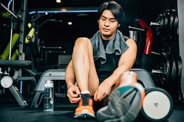 Fototapeta na wymiar A muscular young man is tying his shoelaces on the gym floor, surrounded by dumbbells and other equipment. The shot emphasizes his strength and endurance during exercise