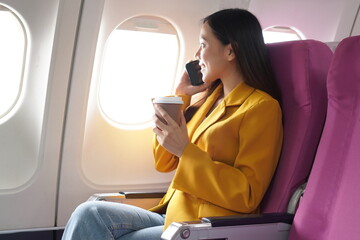 Asian woman sitting on airplane playing with cell phone and looking out window on vacation travel...