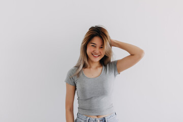 Happy Asian Thai woman wear grey, smiling and raising her hand up for touching and grabbing hair, looking at empty camera, chilling standing isolated on white background wall.