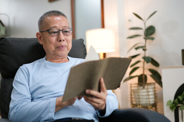 Happy senior Asian man reading book on the chair in living room at home - Powered by Adobe