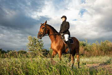 Obraz premium Horsewoman in equestrian sports gear, riding a horse, against an expressive sky, horseback riding in the open air