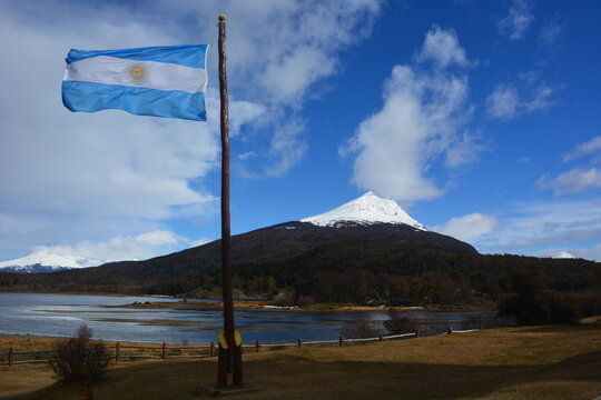 paisaje de cordillera nevada con lago cielo celeste con nubes verticales ,mastil con bandera flameando argentina.