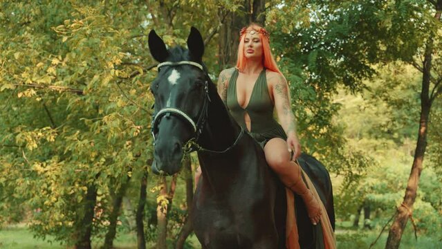 Fashion model posing rides a horse. happy fantasy woman forest nymph hand strokes horse muzzle sitting on horseback. Fairy tale girl elf sexy dress red long hair. summer nature forest green trees. 4k