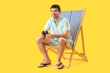 Young man with binoculars sitting in deck chair on yellow background