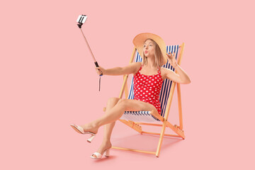 Beautiful young woman taking selfie in deck chair on pink background
