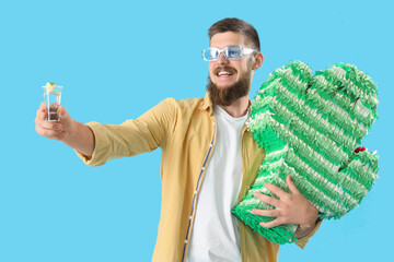 Handsome young man with tequila and pinata on blue background