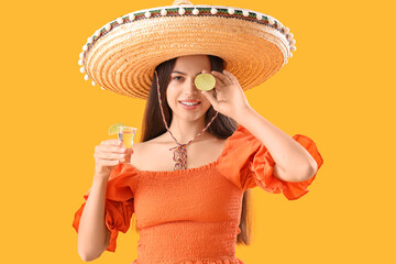 Beautiful young Mexican woman in sombrero and with tequila on yellow background