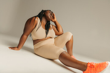 Young plus size African woman in sportswear looking relaxed while sitting against studio background