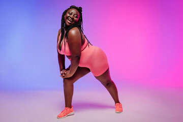 Young voluptuous African woman in sportswear exercising and looking happy on colorful background