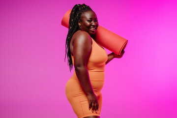 Happy plus size African woman in sportswear carrying exercise mat on pink background