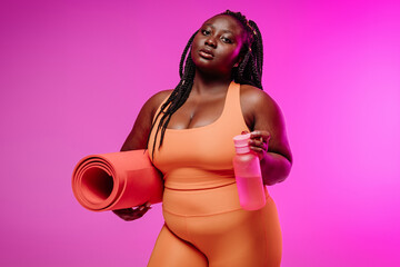 Curvy African woman in sportswear carrying exercise mat and looking confident on pink background
