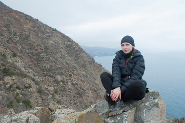 A girl in a jacket and heavy boots sits on the top of a mountain. Hiking in a natural landscape...