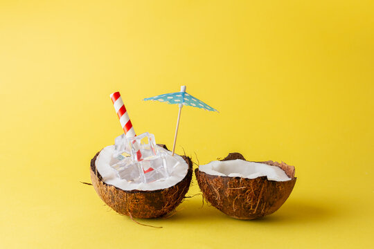 Coconut drink abstract made of coconut with ice cubes, drinking straw and paper umbrella parasol isolated on yellow background.