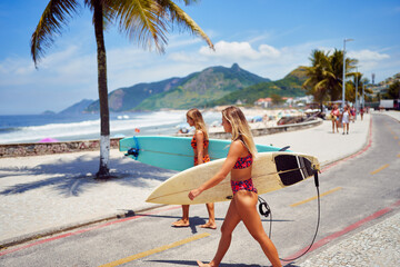 two caucasian women surfers walking on the street holding surfboards in swimsuit 