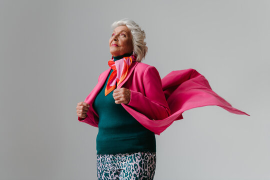 Elegant elderly woman looking like super hero while wearing pink coat against grey background