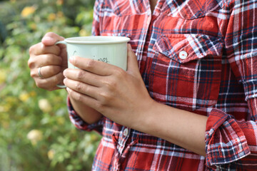 Woman hands holding enamel mug in the park outdoor. Woman wears red plaid shirt.