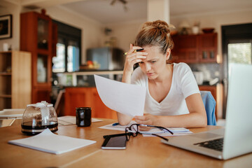 Concerned young woman reading through bank statements at home - obrazy, fototapety, plakaty