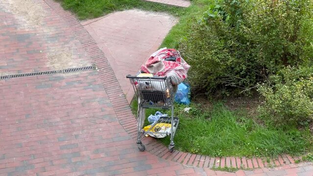 verlassener Einkaufswagen eines Obdachlosen mit M&uuml;ll und Habseligkeiten beladen am Stra&szlig;enrand 