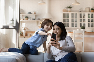 Happy Asian mother and Caucasian son having fun with smartphone, laughing young mom with 7s boy...