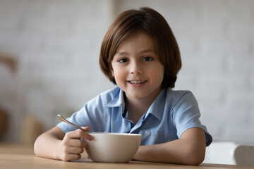 Head shot portrait smiling 8s boy enjoying having breakfast in kitchen, happy adorable child kid looking at camera, sitting at table with tasty cereals or porridge bowl and spoon in morning