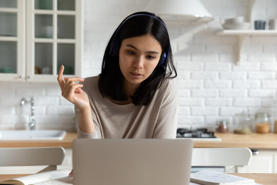 Focused Asian woman in headphones watching webinar, using laptop, studying at home, concentrated young female student preparing to exam, engaged in educational online course, distance education