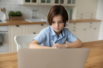 Serious 8s boy in headphones looking at laptop screen, watching webinar, studying online in kitchen at home, focused child student listening to lecture and writing taking notes, homeschooling concept