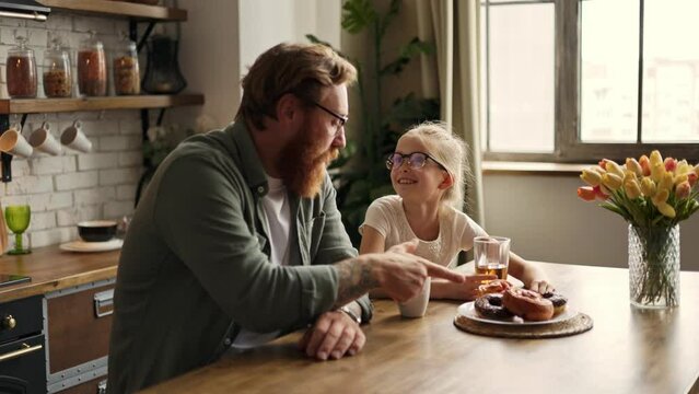 Smiling child and father talking near pastry and drinks in kitchen at home, slow motion