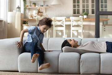 7s boy having fun while smiling Asian mother relaxing lying on comfortable cozy couch at home, happy young mom with Caucasian son spending leisure time in living room together, enjoying weekend