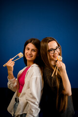 Fototapeta premium Two young women stand with their backs to each other and hold sushi rolls with chopsticks near their mouths