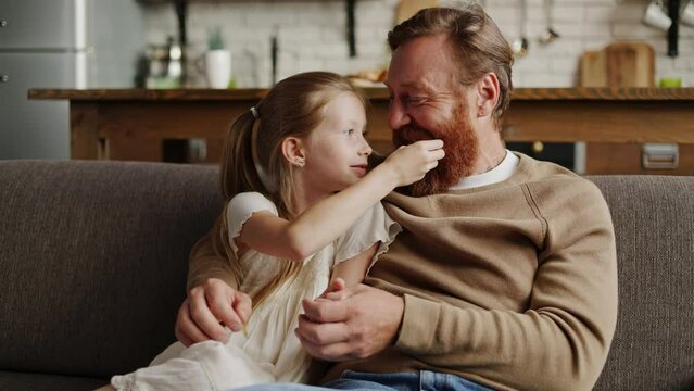 Bearded and redhead father hugging cheerful daughter on couch at home, slow motion