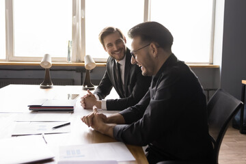 Two overjoyed 35s colleagues, businessmen having fun, enjoying informal conversation at meeting in modern office, laughing at funny jokes, smile, working together at workplace. Teamwork, companionship