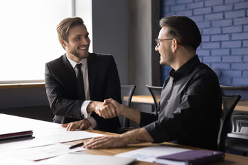 Two satisfied young professional businessmen shaking hands after successful financial deal at meeting. Businesspeople make business agreement in office. Partnership, beneficial cooperation, teamwork