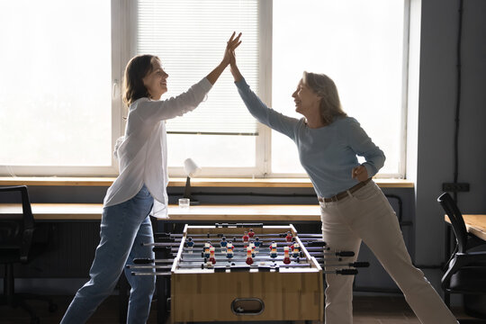 Young and mature women, two office employees playing table football, give high five, celebrate victory feel happy, enjoy lunch break together at workplace. Friendship, fun at workplace, companionship