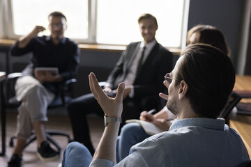Group of office workers sit in circle at meeting in office discuss business idea, engaged in teambuilding activity. Confident male leader explain new strategies to creative team. Teamwork, cooperation