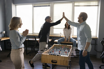 Group of businesspeople rest at lunch break play table foosball, have fun at workplace, enjoy teambuilding activity. Two young man colleagues give high five over board game celebrate final match over - obrazy, fototapety, plakaty