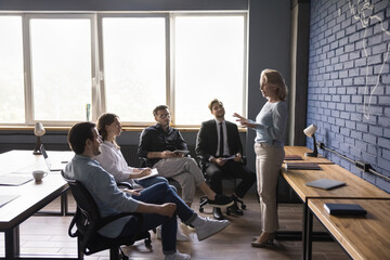 Elderly businesswoman, team leader talk to employees in office, instruct trainees at briefing, offering idea. Young business group listen to mentor speech, discuss work results. Coaching, teambuilding