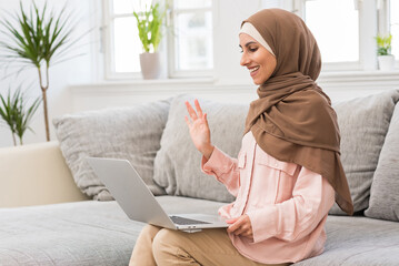 Side view of a beautiful Muslim woman in hijab sitting on a sofa in a bright living room holding...