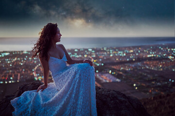artistic portrait beautiful woman with white dress sitting on a rock with stars and milky way over the city in the background