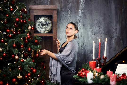 A girl in a black evening dress stands near a large grandfather clock. Festive mood, expectation of a miracle. On the eve of Christmas.