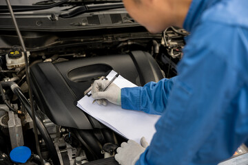Close-up shot of an Asian mechanic's hand, in blue workwear, writing notes on a paper while working on a car in an auto repair shop.