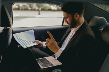 Male entrepreneur sitting on car backseat and working with documents while going to office - obrazy, fototapety, plakaty