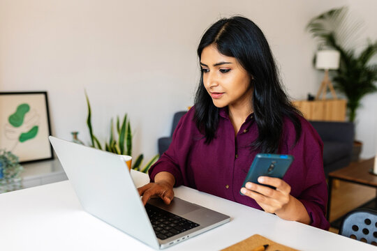 Young indian business woman holding cell phone while typing on laptop keyboard at home. Entrepreneur people, home office and technology lifestyle concept.