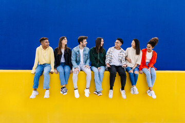 Young group of diverse friends having fun while hanging out sitting on yellow wall over blue...