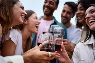 Group multiracial laughing friends toasting glasses red wine and celebrating party outdoors. Young people together cheers on open air. Boys and girls enjoying free time on summer weekend vacation. - obrazy, fototapety, plakaty