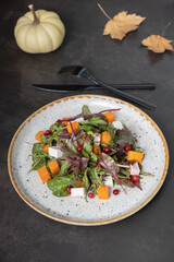 Plate with salad with greens, pumpkin and soft cheese and cutlery on dark background.