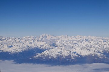 Fototapeta na wymiar Beautiful aerial view of alpine snowcapped mountain range peaking through heavy clouds. Mountain peaks of Italian alps from above. The impressive winter view is taken from an airplane window.